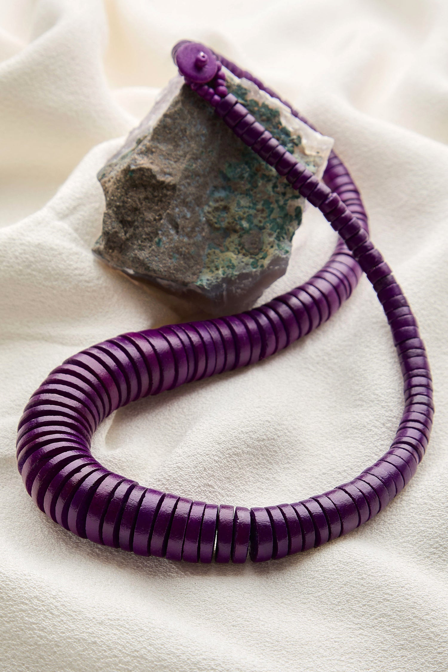 A handmade Short Graduated Coco Disc Necklace with purple segments is coiled and resting against a rough rock atop soft, light-colored fabric.