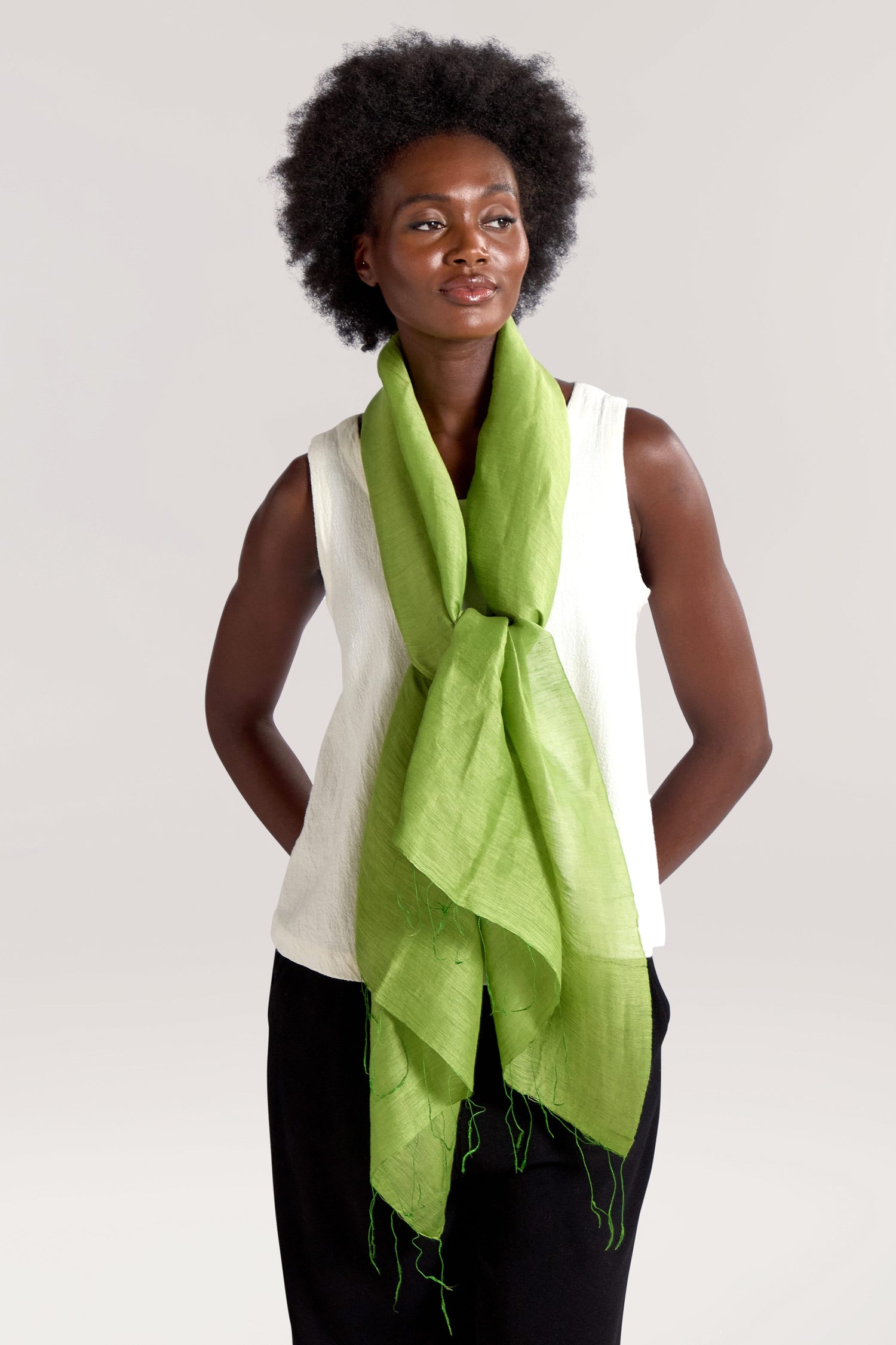 A person with short curly hair, wearing a white sleeveless top and a bright green Soft Linen Scarf draped around their neck, is standing with hands behind their back against a plain background.