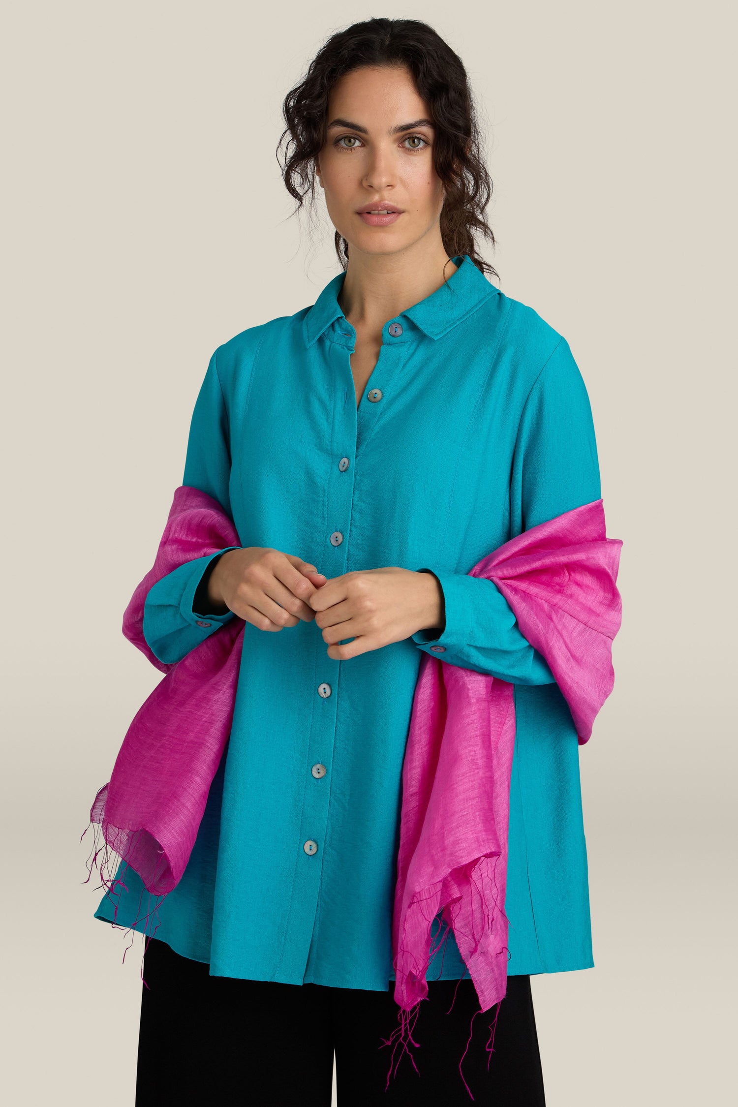 A woman stands against a plain background wearing a teal button-up shirt, holding a Soft Linen Scarf. The bright pink, lightweight scarf is draped elegantly over her arms.