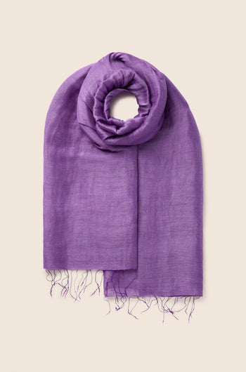 A folded Soft Linen Scarf in a rich deep purple color, adorned with fringe at the ends and showcased against a simple beige background. This exquisite linen accessory is a testament to fair trade craftsmanship.