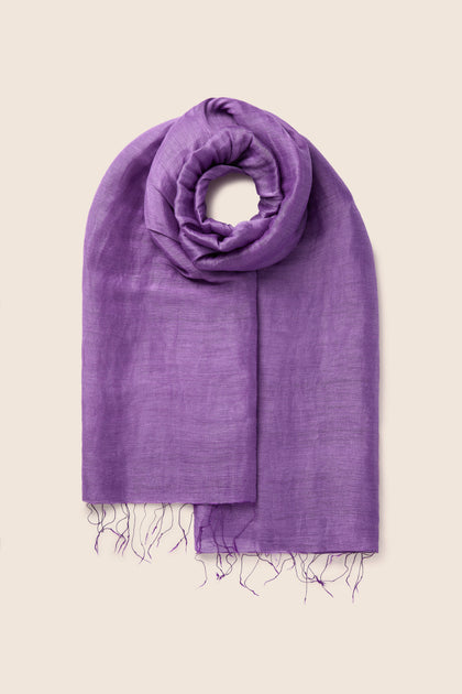 A folded Soft Linen Scarf in a rich deep purple color, adorned with fringe at the ends and showcased against a simple beige background. This exquisite linen accessory is a testament to fair trade craftsmanship.