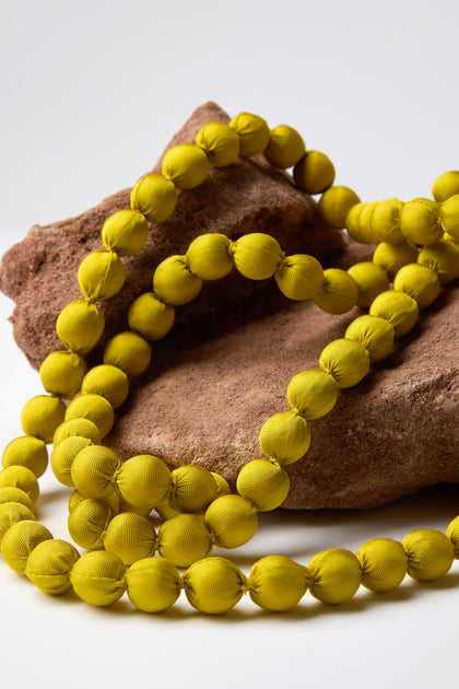 A Handmade Long Silk Spheres Necklace, displaying vibrant colors, is elegantly arranged over two uniquely shaped brown stones on a simple white backdrop.