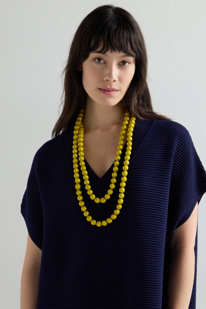 A person with long dark hair wearing a navy textured top and layered Handmade Long Silk Spheres Necklace, highlighting artisanal craftsmanship, stands against a plain light background.