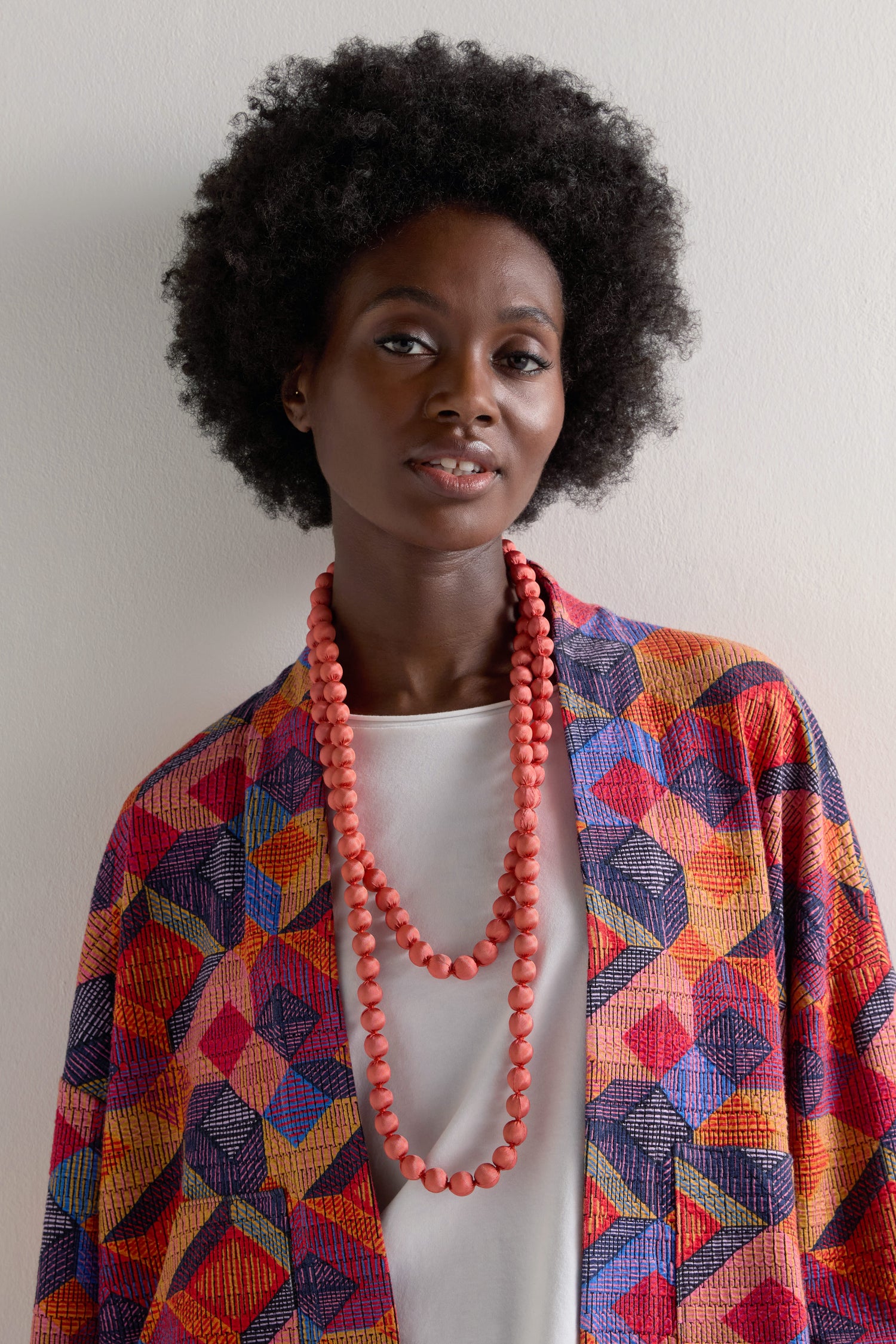 A person with an afro hairstyle wears a vibrant geometric patterned garment and accessories, including a Handmade Long Silk Spheres Necklace and layered coral pieces, against a plain background.