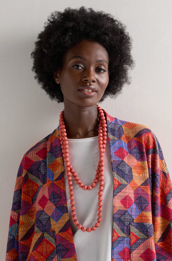 A person with an afro hairstyle wears a vibrant geometric patterned garment and accessories, including a Handmade Long Silk Spheres Necklace and layered coral pieces, against a plain background.