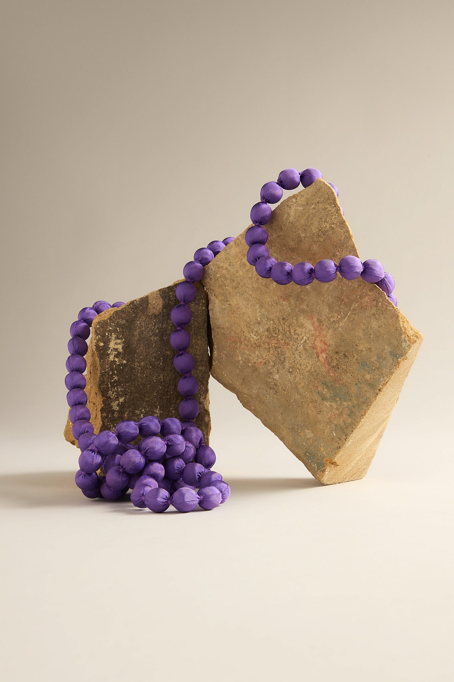 A Handmade Long Silk Spheres Necklace, featuring artisanal craftsmanship with purple beads, drapes elegantly over two uniquely shaped rocks against a neutral background.