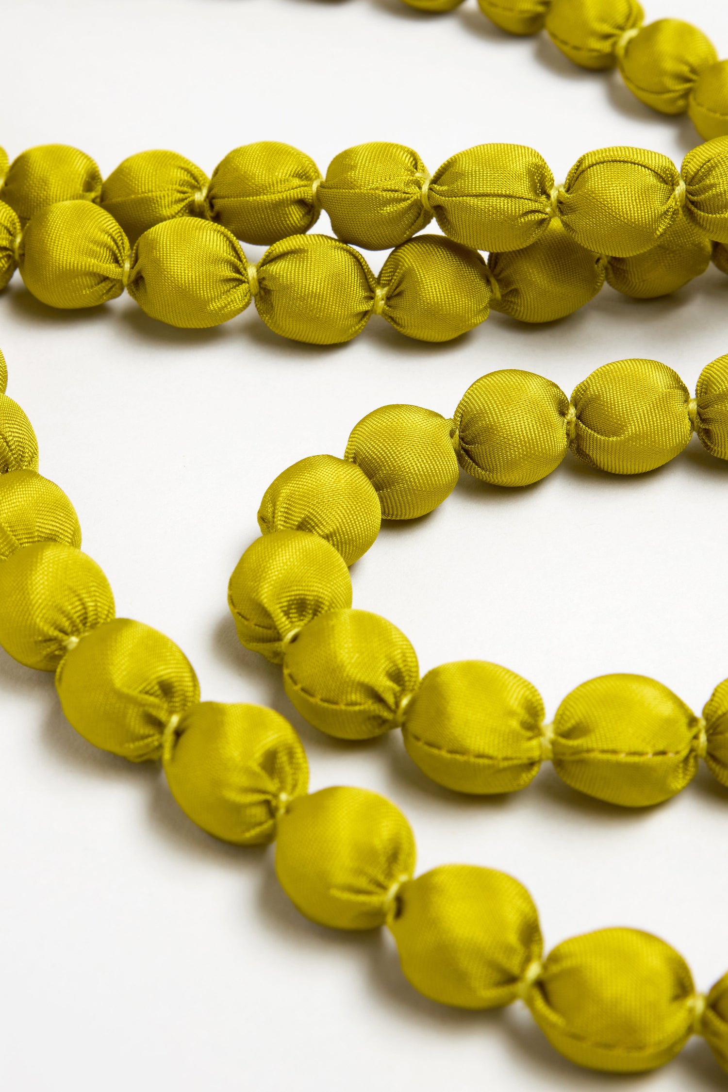 Close-up of the Handmade Long Silk Spheres Necklace featuring bright yellow-green, fabric-covered beads arranged in curved lines on a white surface, highlighting its artisanal style and the beauty of handmade silk jewelry.