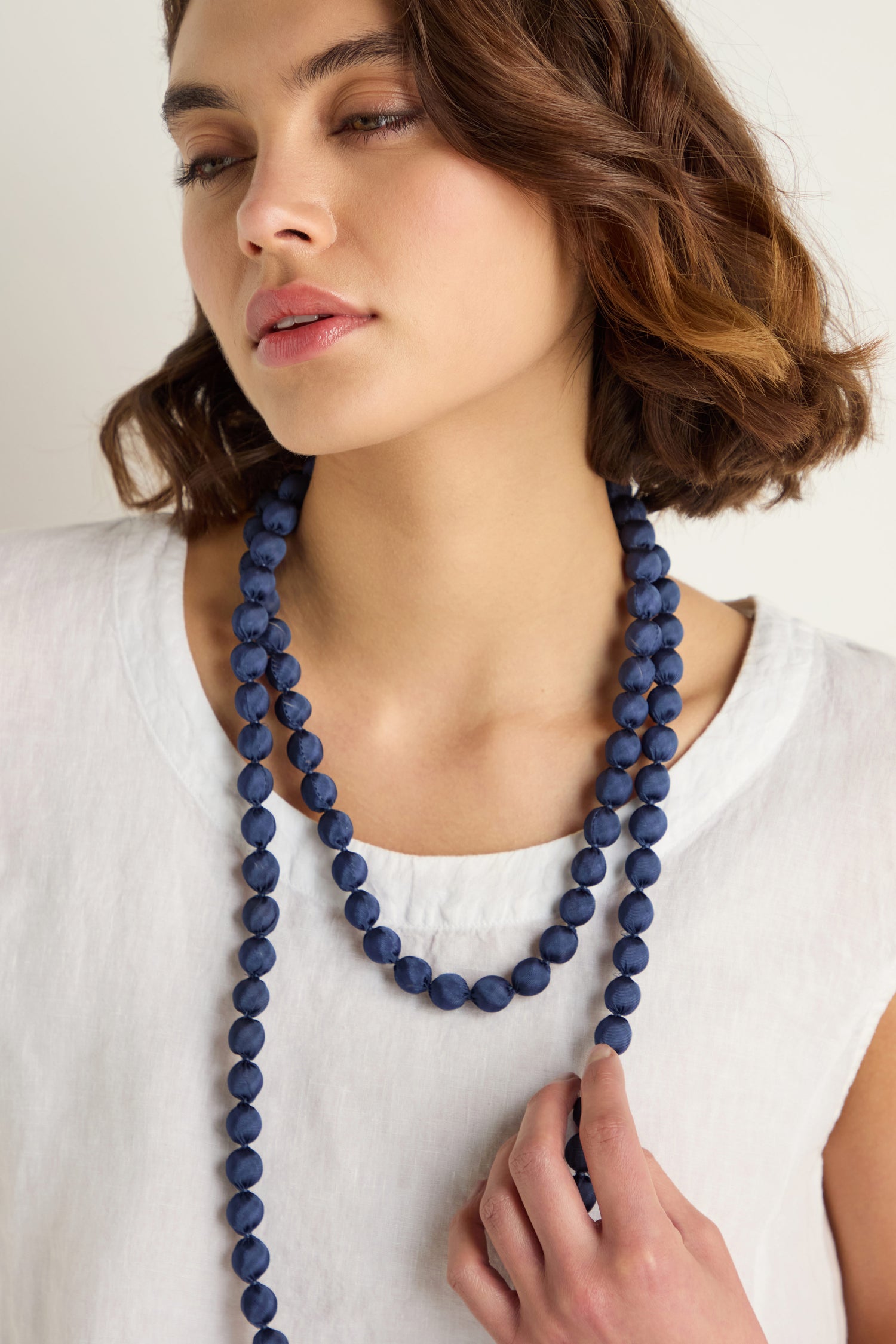 A woman with short brown hair, wearing a white top, showcases two long dark blue Handmade Long Silk Spheres Necklaces, one hand resting gently on her chest.