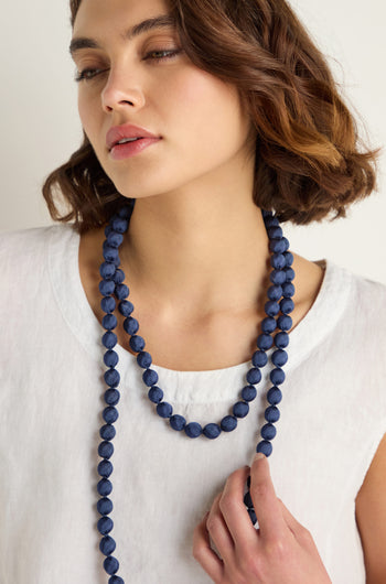A woman with short brown hair, wearing a white top, showcases two long dark blue Handmade Long Silk Spheres Necklaces, one hand resting gently on her chest.
