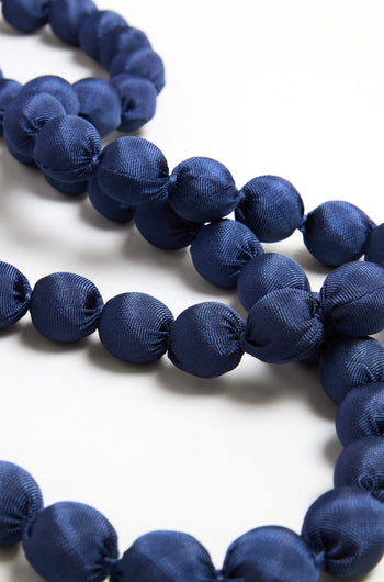 A close-up of the Handmade Long Silk Spheres Necklace showcases large, round, navy blue silk-wrapped beads arranged in strands on a white surface.