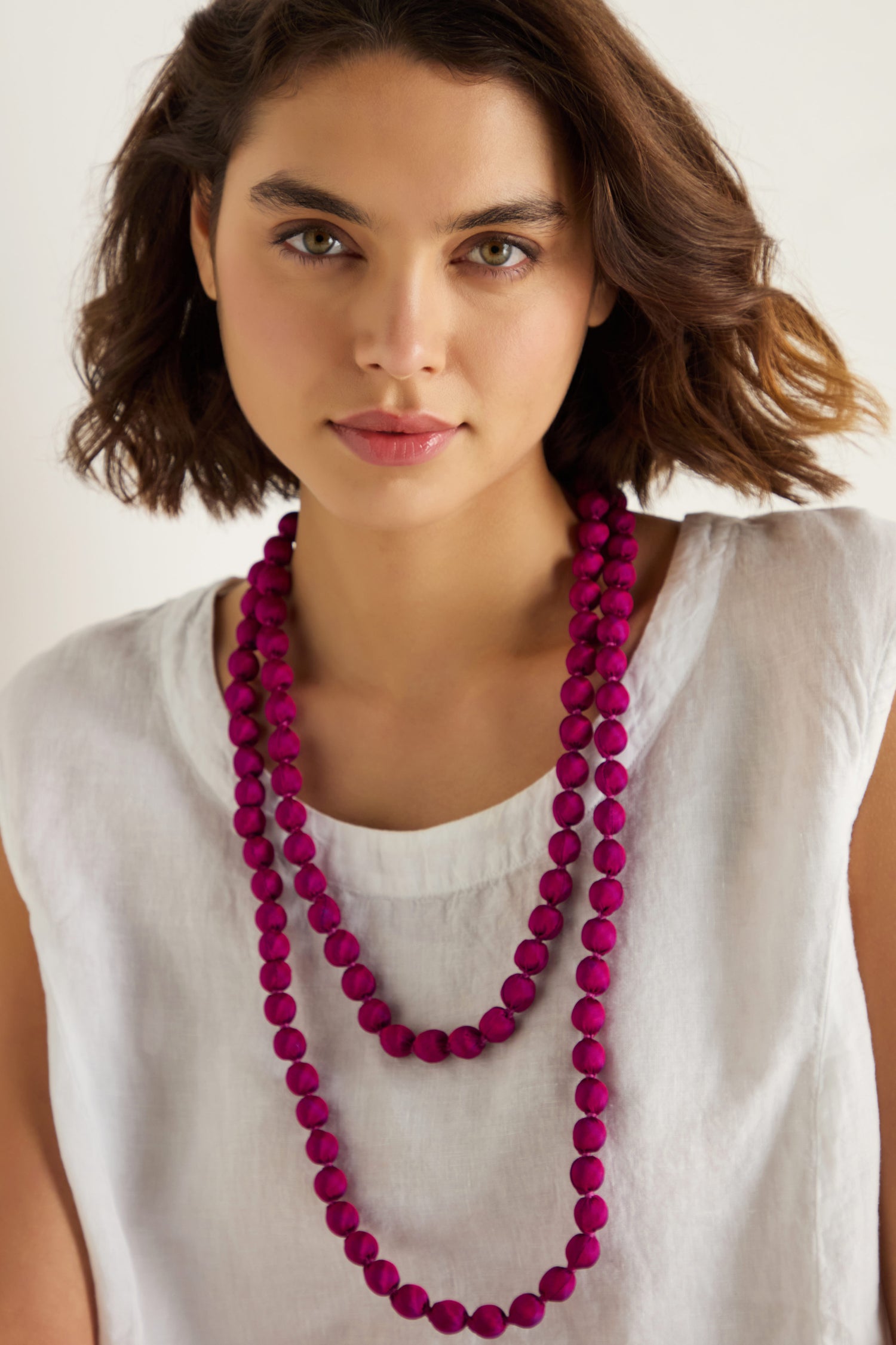A woman with short brown hair wears a white top and two Handmade Long Silk Spheres Fuchsia Necklaces, each artisan-made and layered with magenta beads.