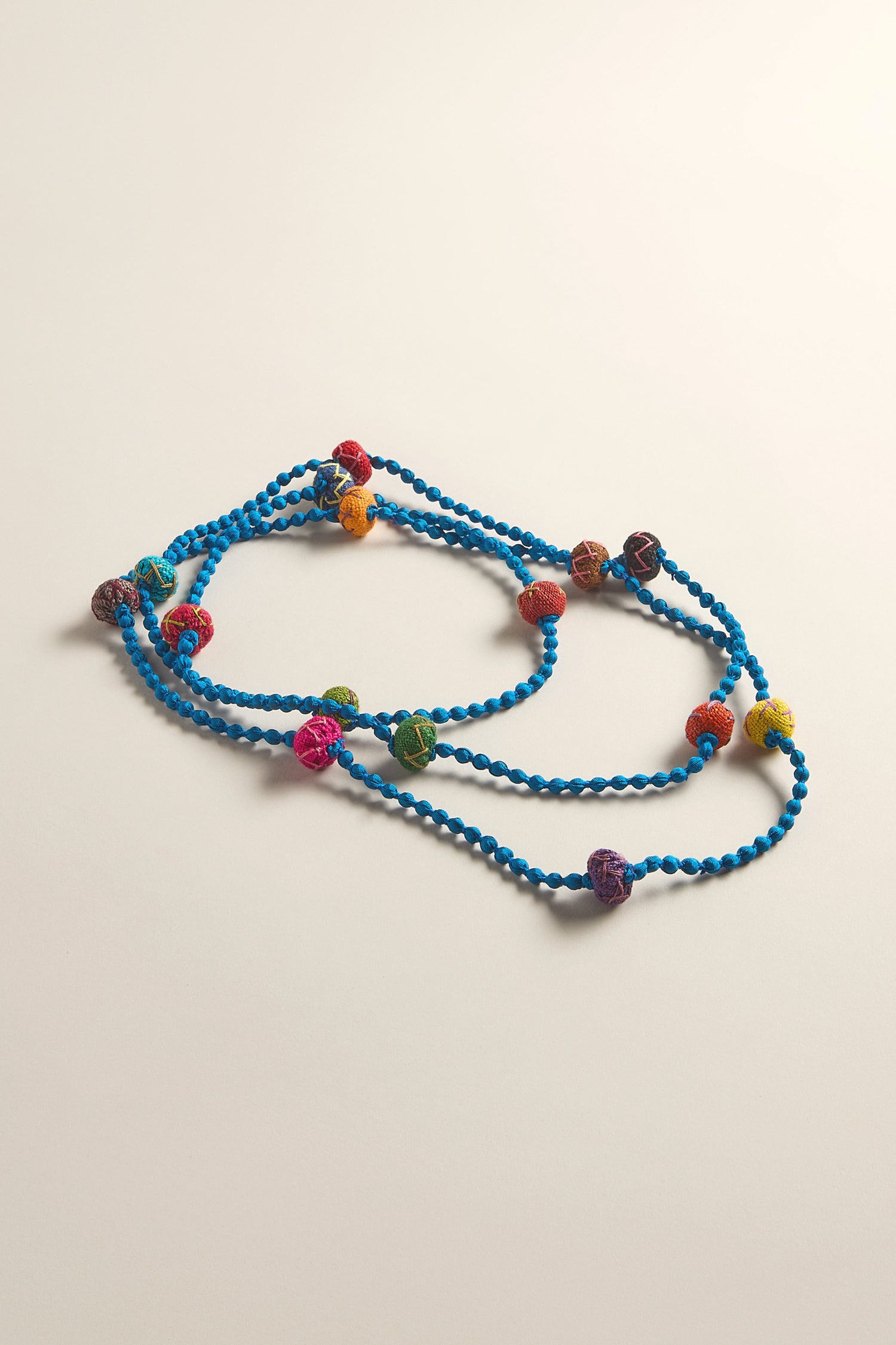 The Handmade Rainbow Silk Spheres Necklace, with a blue beaded strand and colorful crocheted silk spheres, is elegantly coiled on a plain surface.