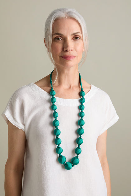 A woman with gray hair is wearing a white blouse and a long Handmade Silk Spheres Necklace showcasing vibrant turquoise hues. She is standing against a neutral background.