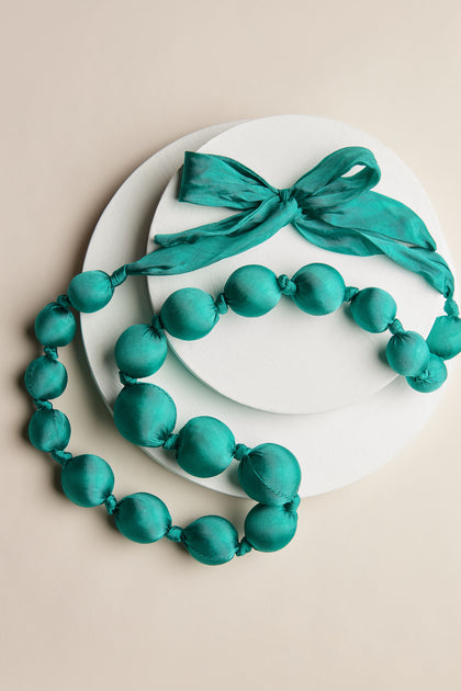 A Handmade Silk Spheres Necklace in vibrant hues of teal, complemented by a matching ribbon, is elegantly displayed on a white round backdrop. The artisanal craftsmanship shines through in every detail.