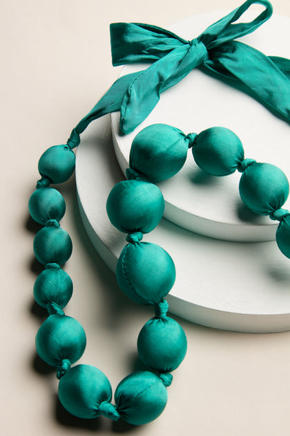 A Handmade Silk Spheres Necklace in a stunning teal hue, adorned with matching bows and showcasing artisanal craftsmanship, is elegantly displayed on two round white stands.