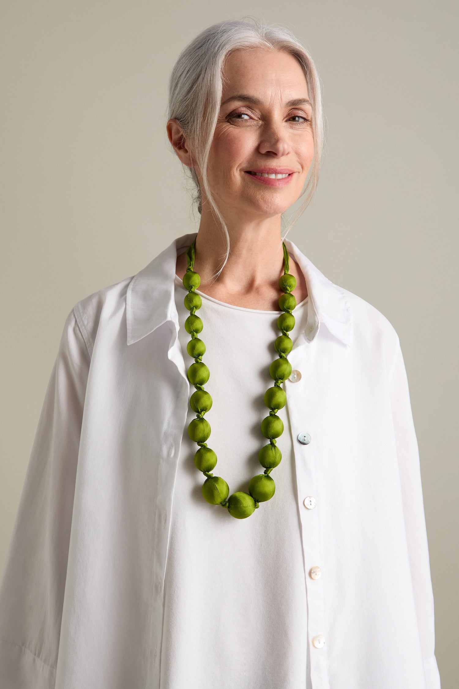 A woman with grey hair wearing a white shirt smiles, showcasing a Handmade Silk Spheres Necklace that highlights artisanal craftsmanship.