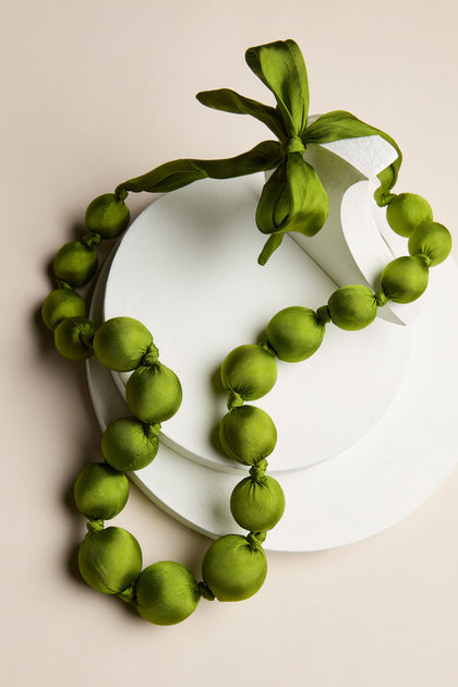 The Handmade Silk Spheres Necklace, adorned with green fabric-covered beads and a tied ribbon, is elegantly displayed on white circular platforms against a beige background, highlighting its vibrant colors and exquisite artisanal craftsmanship.