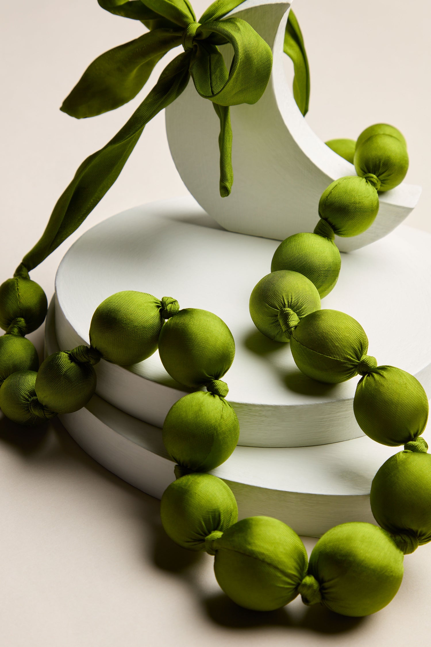 The Handmade Silk Spheres Necklace, adorned with handmade silk spheres in vibrant hues of green and complemented by a green fabric bow, is elegantly displayed on a white, tiered stand.