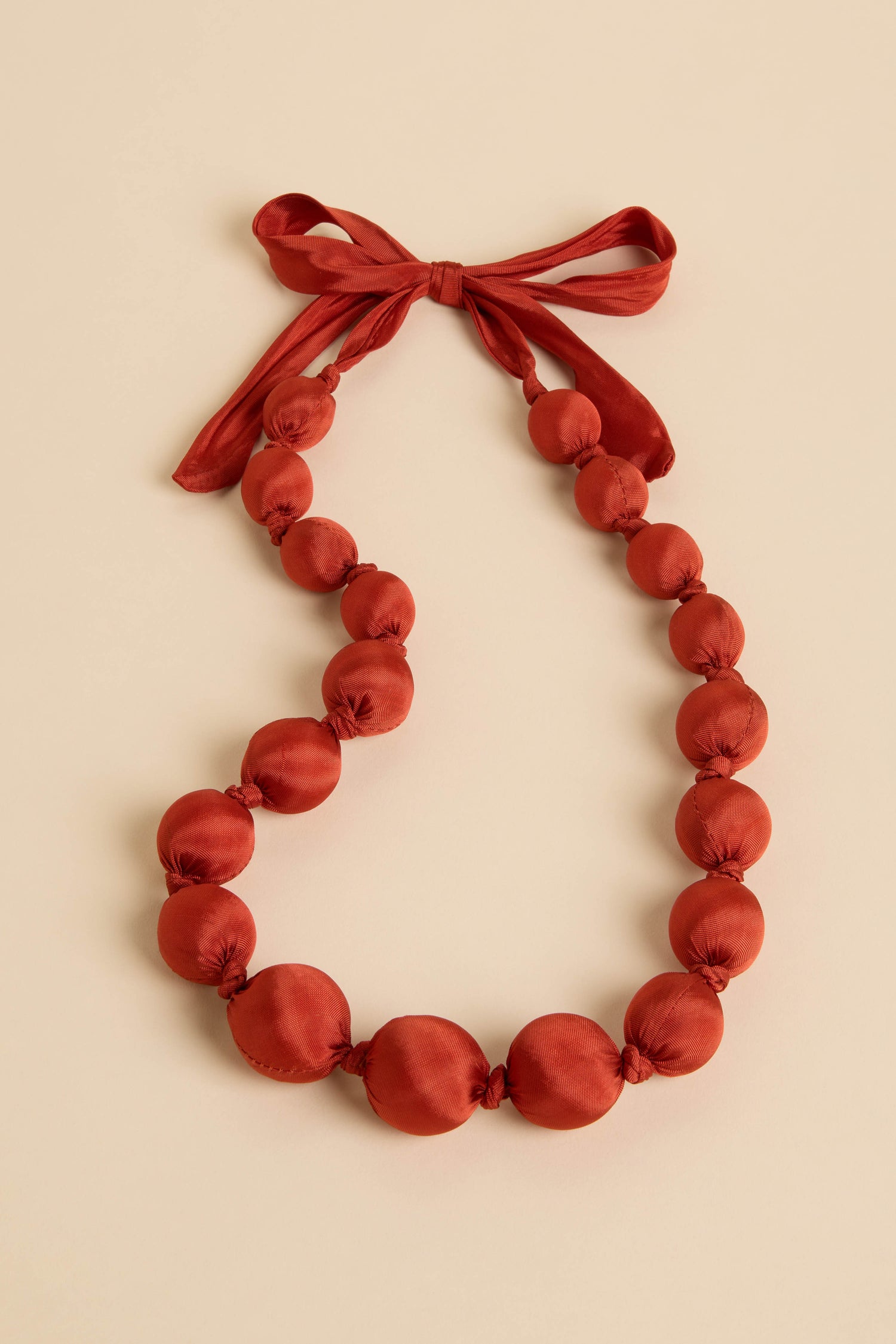 Handmade Silk Spheres Necklace features red, round silk beads evenly spaced with a ribbon bow, highlighting artisanal craftsmanship against a neutral background.
