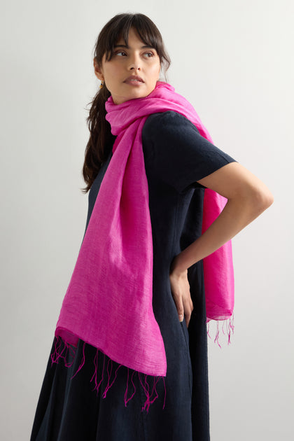 A woman in a black dress is elegantly styled with the Soft Linen Fringe Scarf, a bright pink piece draped over her shoulders. This bohemian accessory adds flair as she gazes to the side against a neutral background.