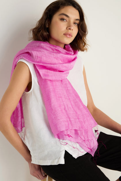 Wearing a sleeveless white top, black pants, and the Soft Linen Fringe Scarf in bright pink, a person sits against a plain light background.