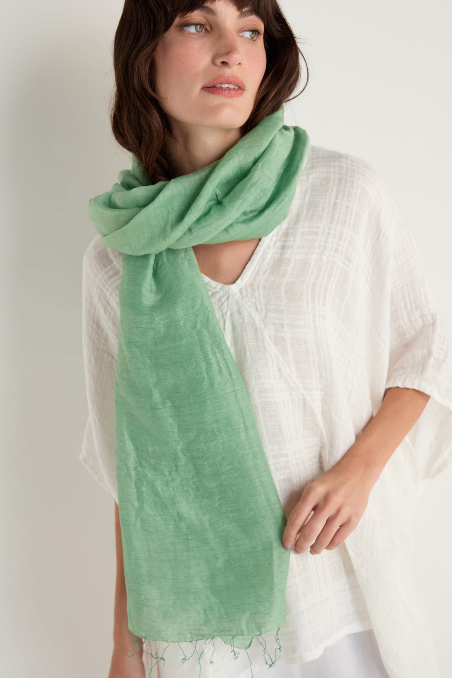 A person stands against a plain light background, wearing a textured white top and an Olive Soft Linen Fringe Scarf in light green, draped around their neck.