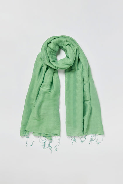 The Olive Soft Linen Fringe Scarf is a lightweight, light green accessory with a slightly crinkled texture and fringed ends, crafted from a soft linen-silk blend and shown laid flat on a white background.