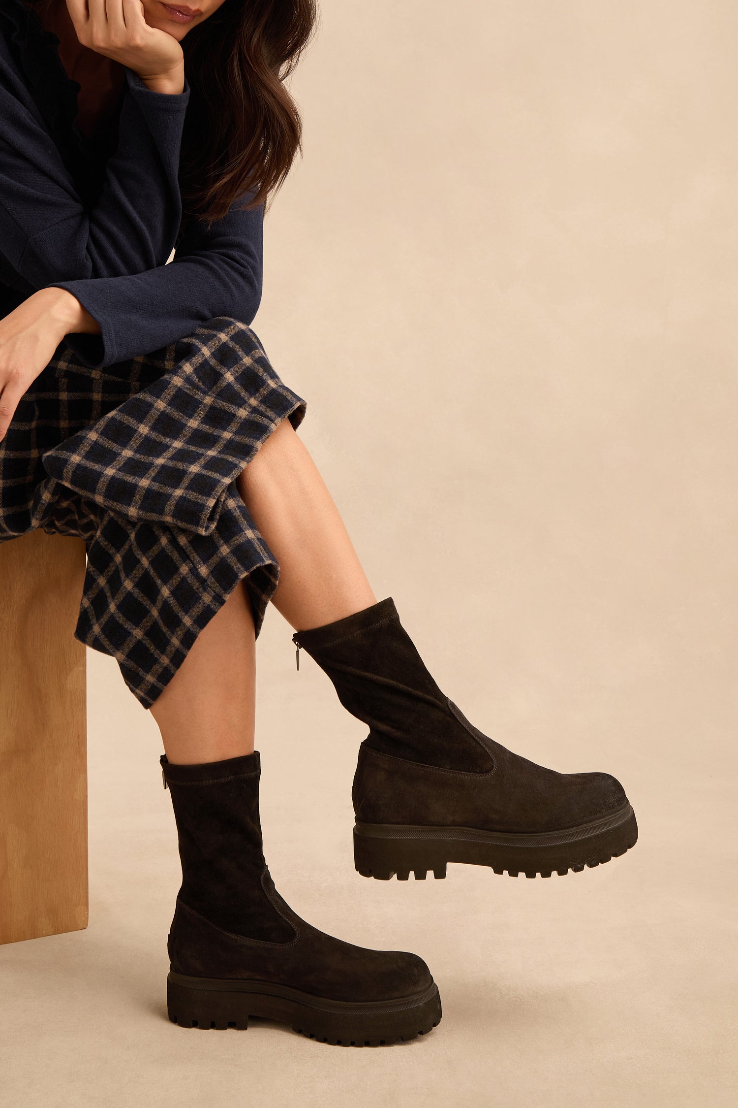 A person sits on a wooden bench wearing a navy plaid skirt, a dark blue top, and Short Stretch Suede Boots with chunky soles, with one hand resting on their chin.