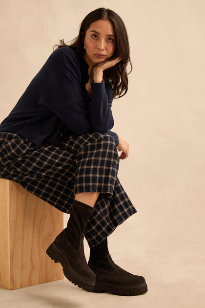 A woman sits on a wooden block, resting her chin on her hand. She wears a navy sweater, plaid pants, and stylish Short Stretch Suede Boots with chunky soles against a neutral background.