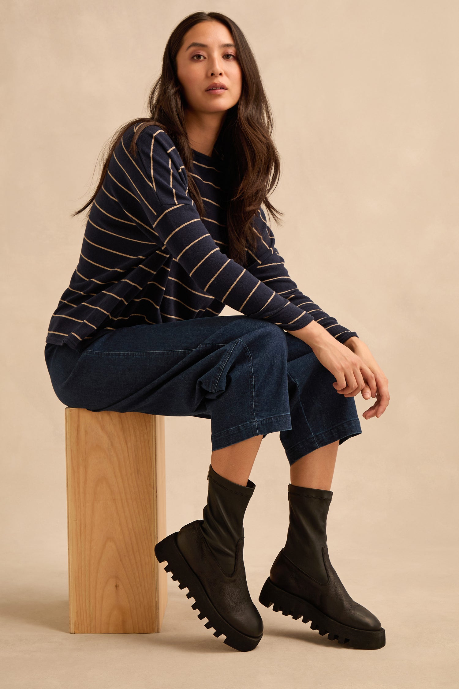A woman sits on a wooden block against a plain beige background, wearing a striped long-sleeve shirt, blue cropped pants, and Platform Napa Leather Stretch Boots with a cleated platform sole.