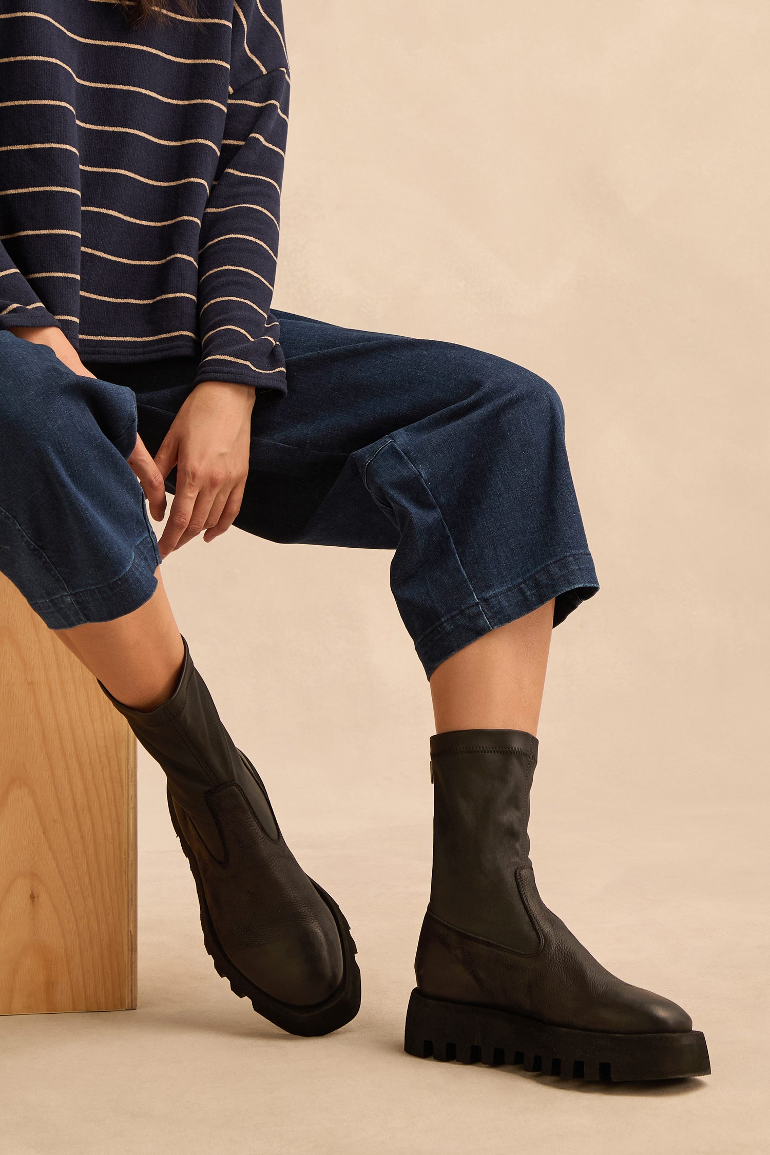 Seated on a wooden block, the person wears a navy striped top, wide-leg jeans, and Platform Napa Leather Stretch Boots featuring a black stretch design and a cleated platform sole against a neutral background.