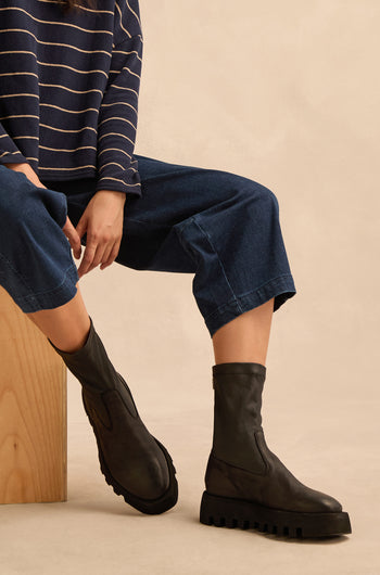 Seated on a wooden block, the person wears a navy striped top, wide-leg jeans, and Platform Napa Leather Stretch Boots featuring a black stretch design and a cleated platform sole against a neutral background.