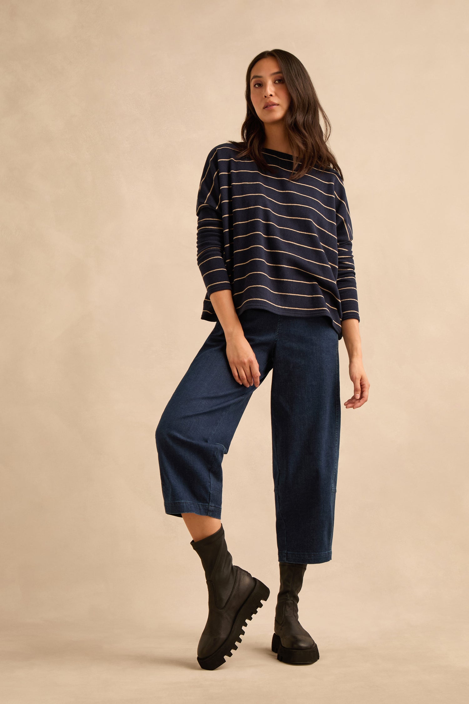 A woman stands against a beige studio backdrop wearing a navy striped long-sleeve top, wide-leg cropped denim pants, and Platform Napa Leather Stretch Boots with a cleated platform sole.