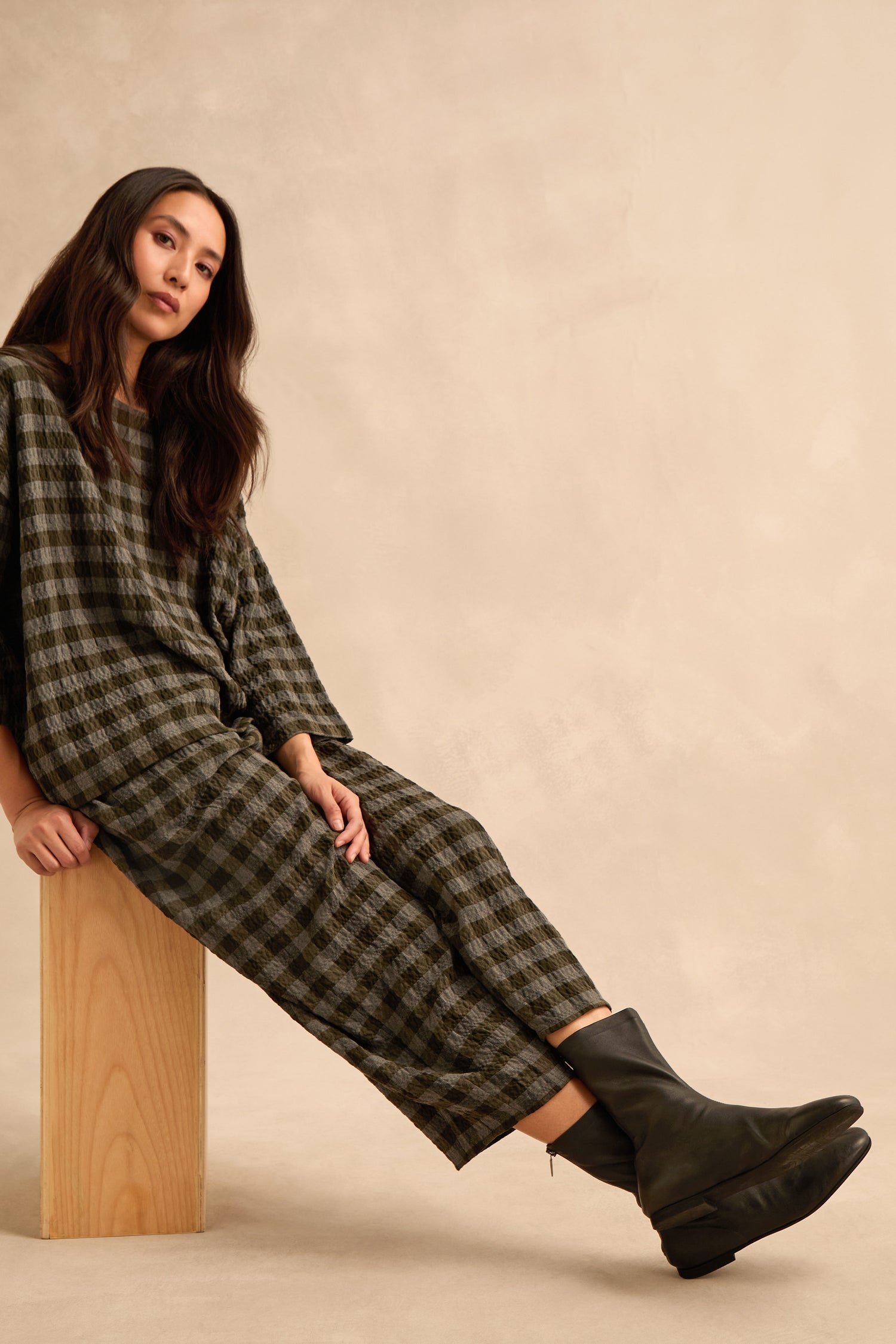 A woman with long dark hair sits on a wooden block, dressed in a checkered outfit and wearing sleek Slim Napa Leather Stretch Boots by a Danish designer, against a neutral beige background.