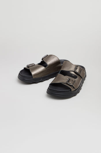 The Double Buckle Leather Sliders feature metallic bronze leather, two adjustable buckles, thick black soles, and are handcrafted in Italy, shown on a plain light background.