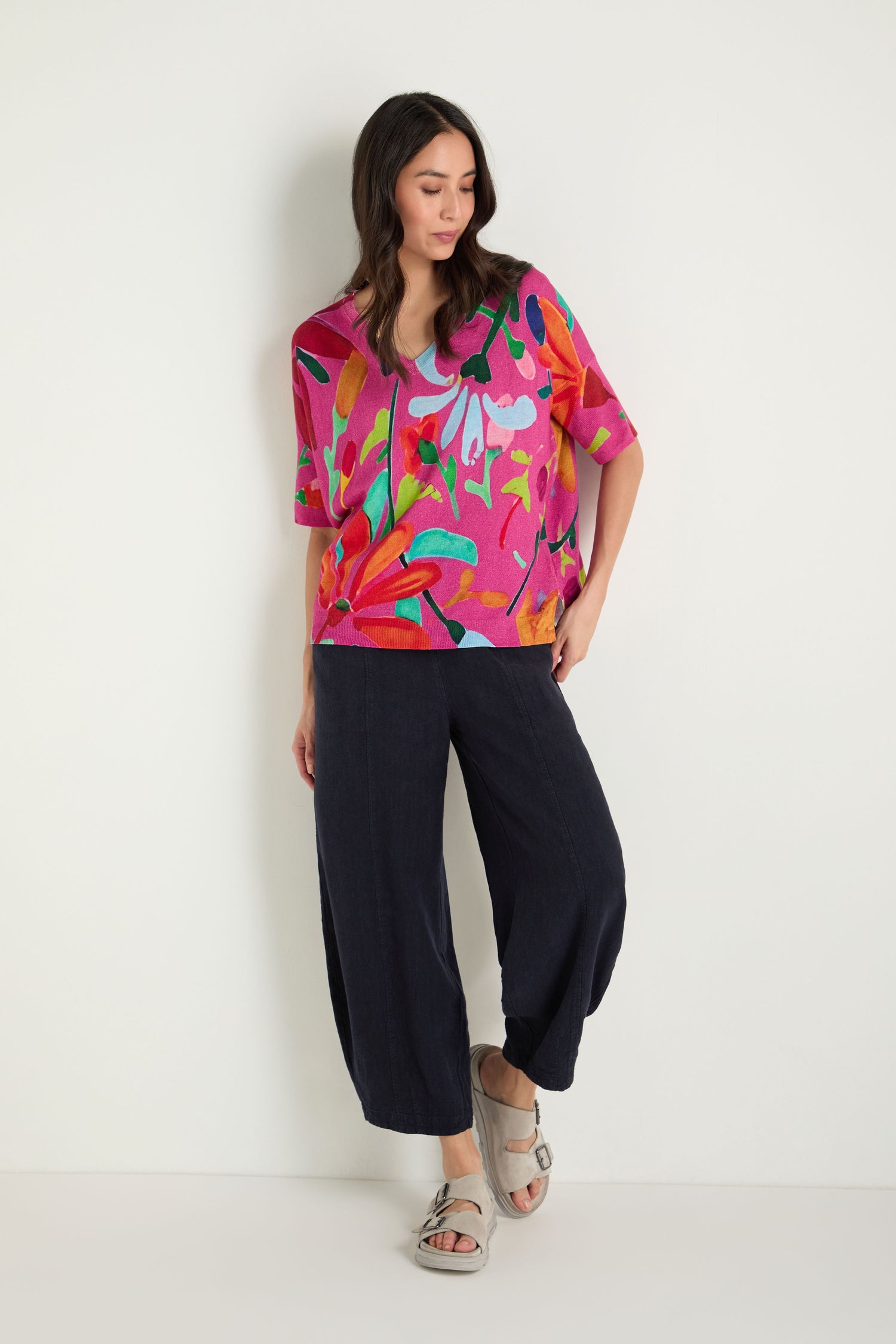 A woman stands against a white wall, one hand in her pocket and looking to the side, wearing a bright floral pink top, loose dark pants, and chic Double Buckle Leather Sliders.