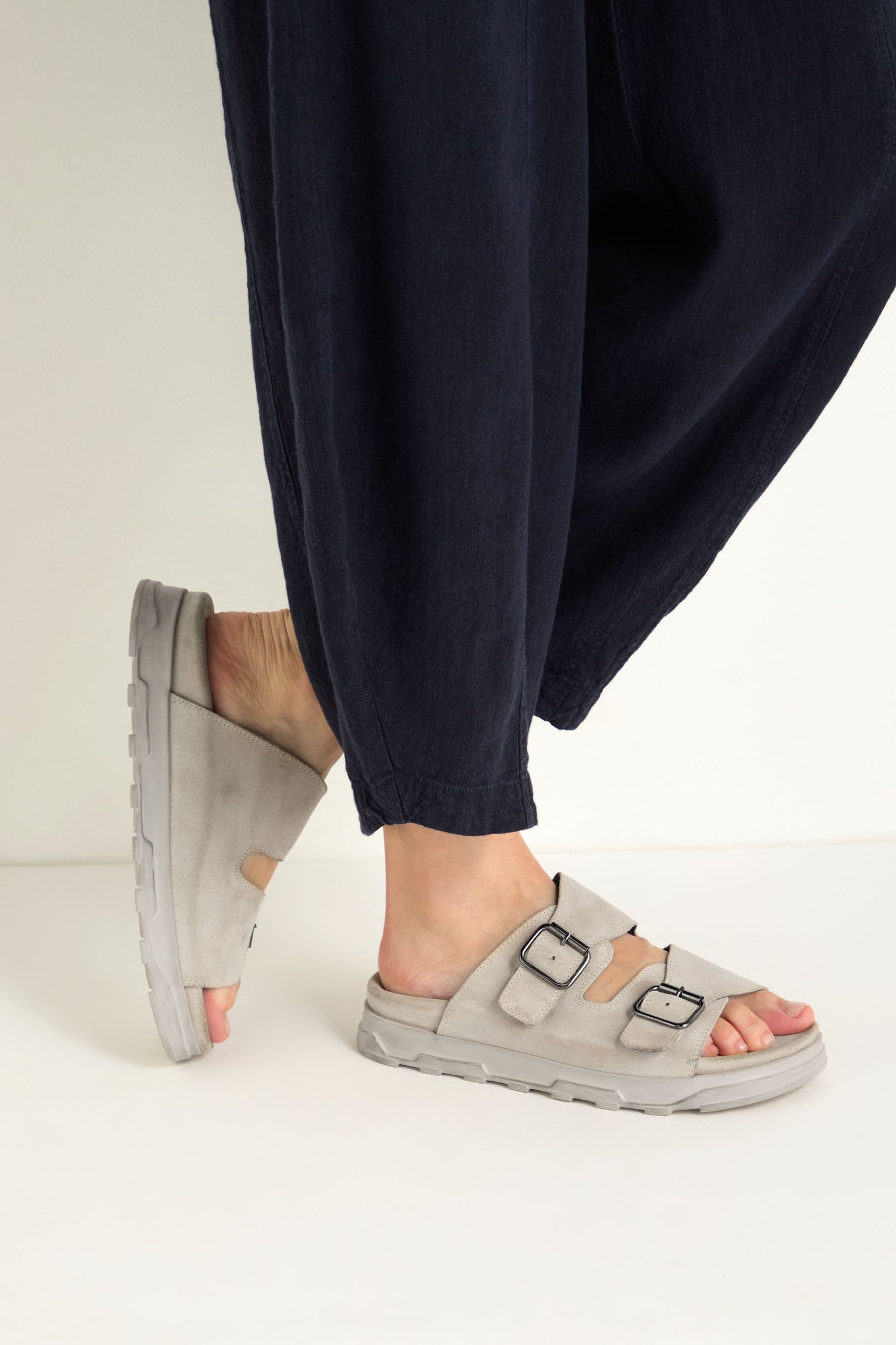 A person stands on a white surface with one foot slightly raised, wearing loose dark pants and light gray Double Buckle Leather Sliders.