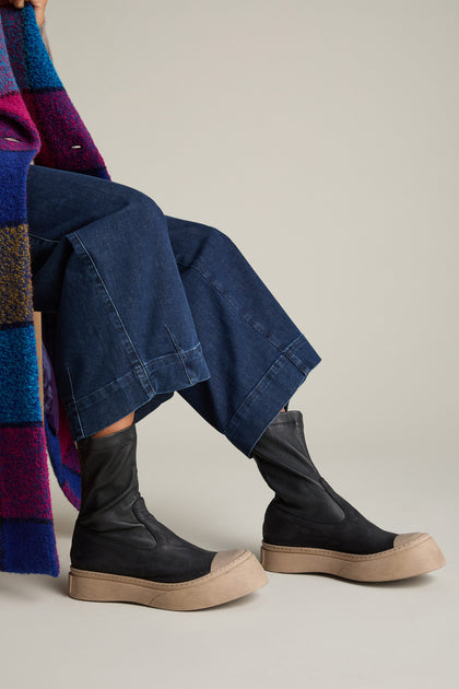 The image shows a person wearing dark blue denim pants, a colorful coat, and black high-top sneakers with thick, light tan soles by Lofina, known as the Contrast Leather Stretch Boot.