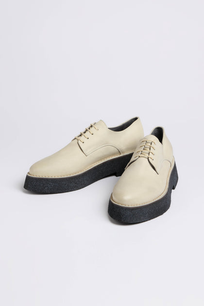 These cream-colored lace-up shoes, handcrafted in Italy, feature thick black soles on a white background. The Lace Up Leather Brogues boast a contemporary platform sole that seamlessly blends style with sophistication.