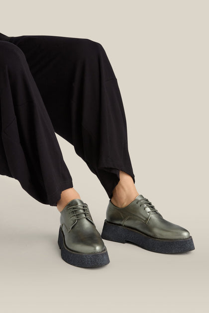 A person seated with both feet visible, dressed in wide-legged black pants, is wearing Lace up Leather Brogues, showcased in a striking shade of metallic grey and featuring a lightweight platform sole.