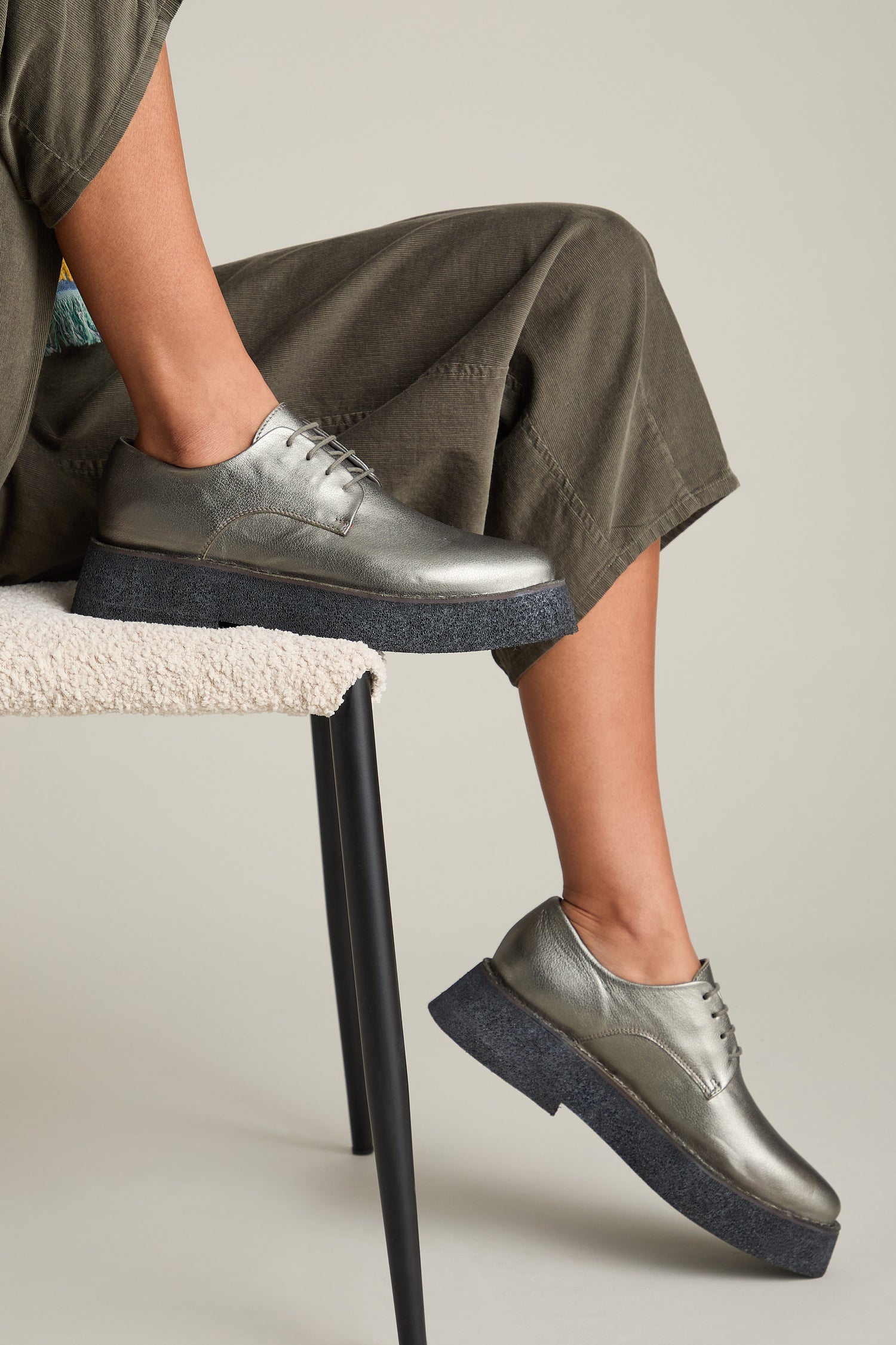 Person wearing chic Lace up Leather Brogues in metallic grey with a lightweight platform sole, sitting on a chair with legs crossed. The person is dressed in grayish-green pants.