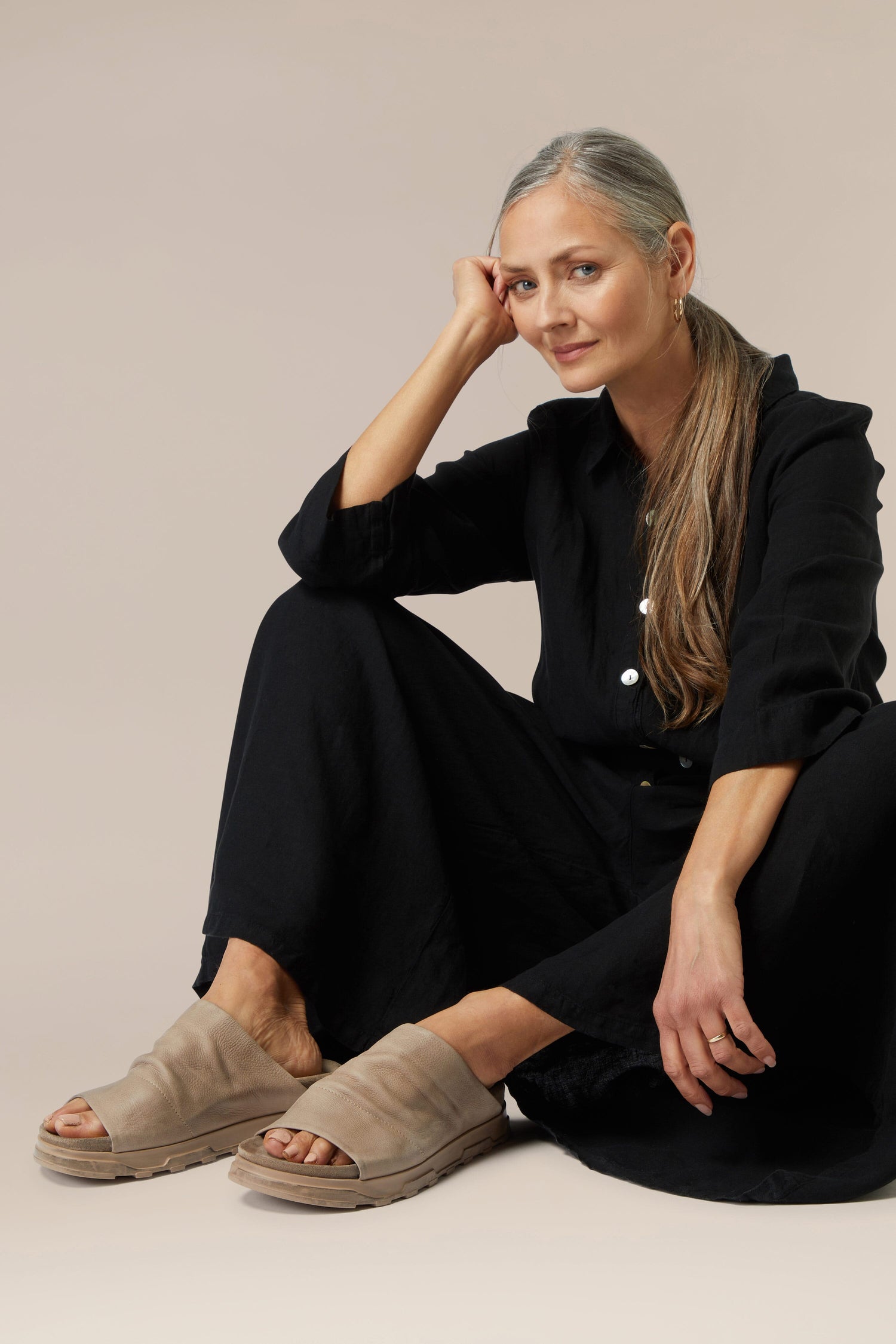 A woman sitting on the floor in a black outfit with a Crushed Leather Slider, resting her head on her hand while looking at the camera.