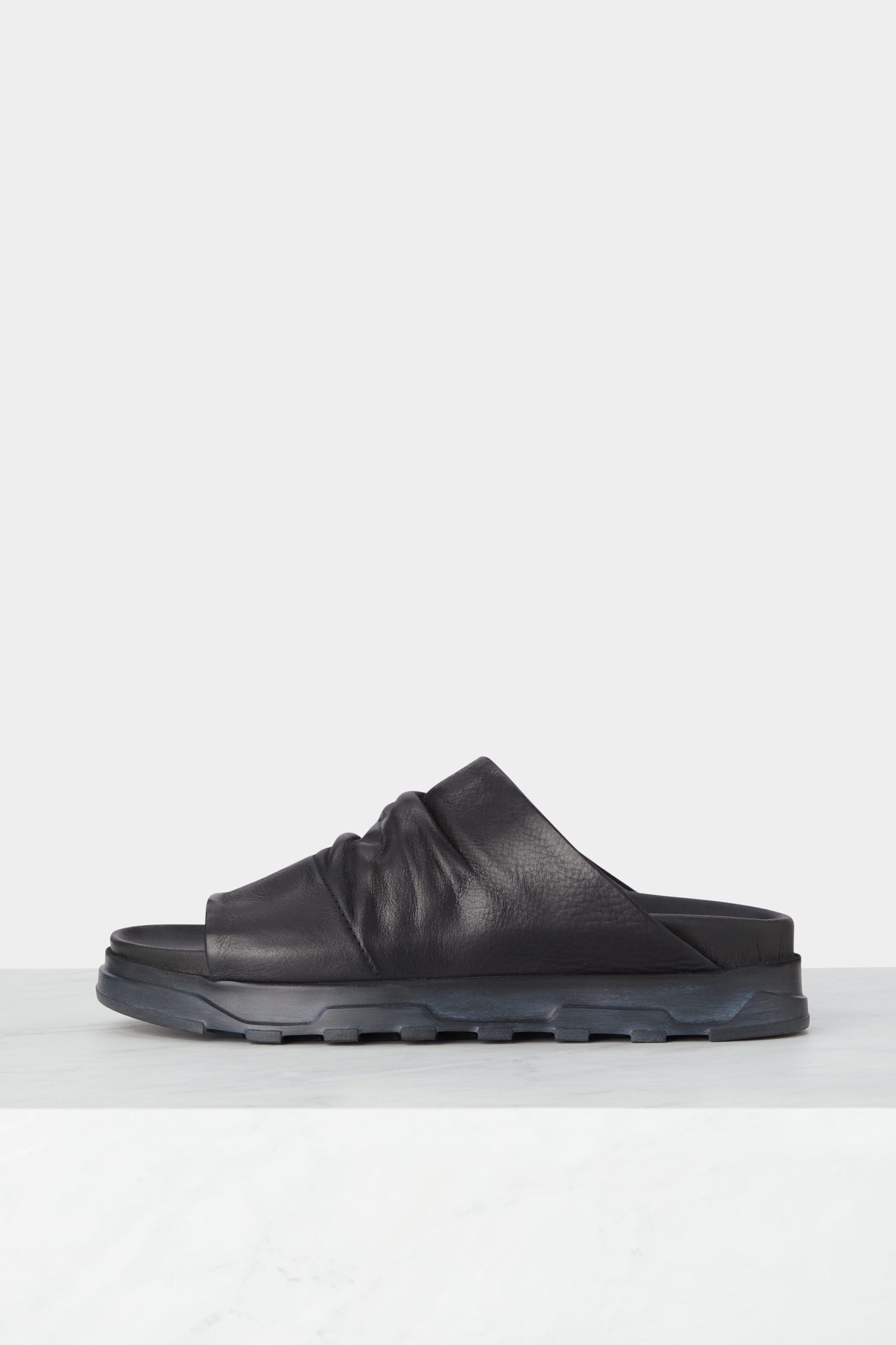 A black Crushed Leather Slider with a ruched leather design and a thick, textured sole is displayed on a plain white surface against a light background, embodying the minimalist elegance of Scandinavian design.