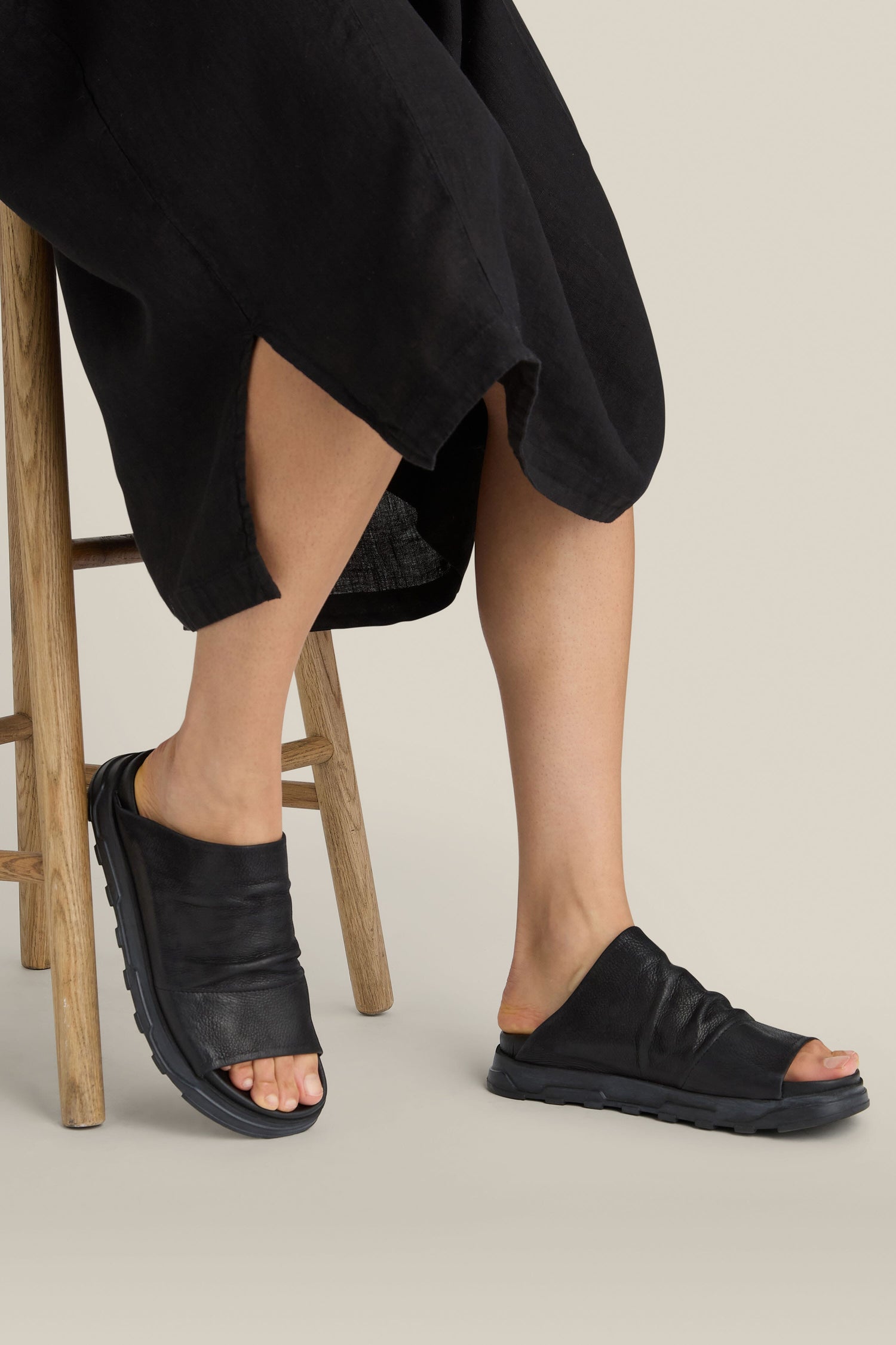 Person seated on a wooden chair wearing a black dress and Crushed Leather Sliders with handmade soles, feet resting on the ground.