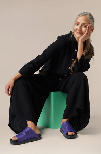 A woman in a black outfit sitting on a green stool against a neutral background, with one hand resting on her cheek and wearing Crushed Leather Sliders.