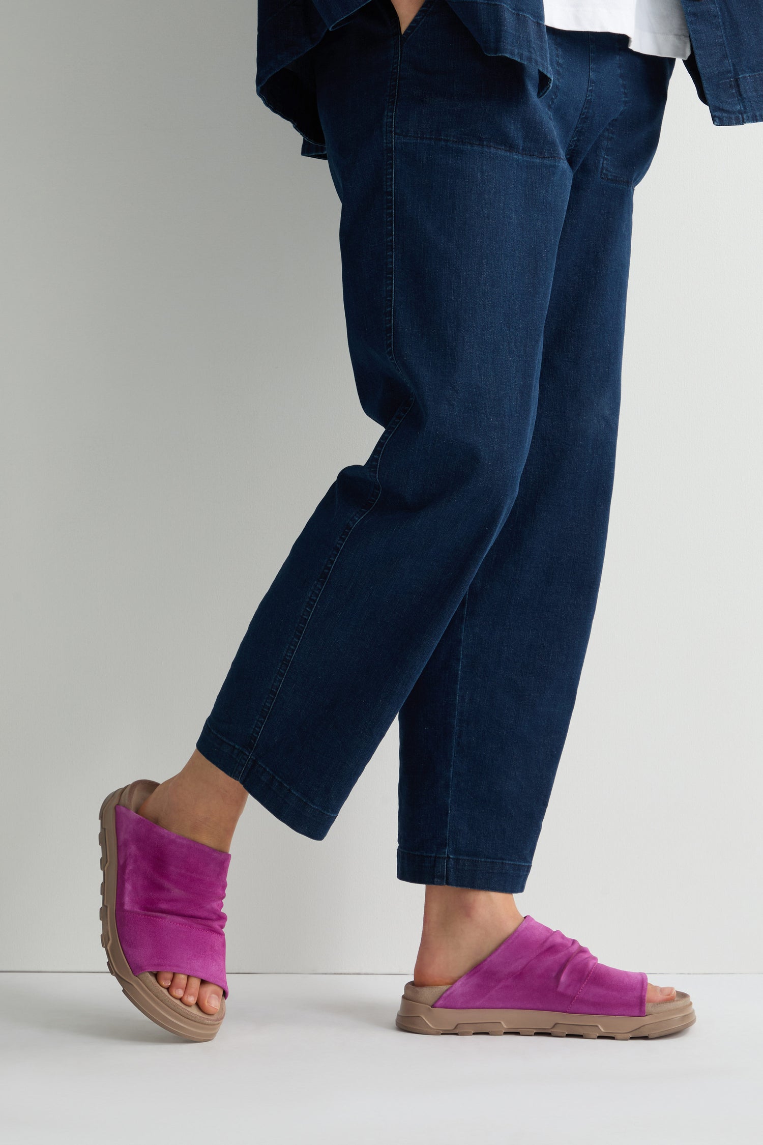 A person stands against a light background, wearing blue denim pants and Magenta Crushed Leather Sliders, radiating all-day comfort.