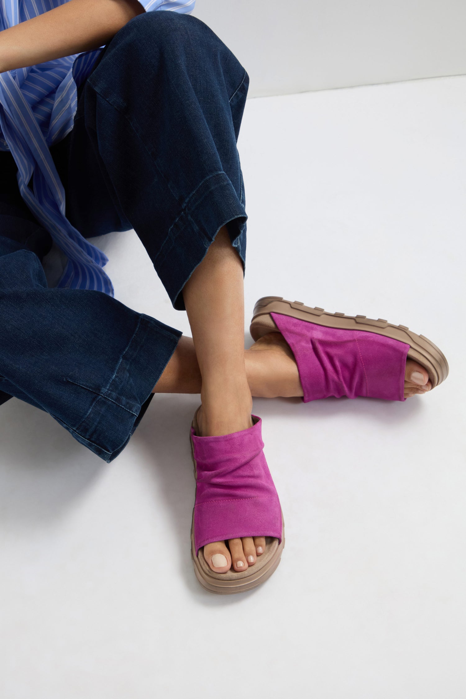 A person seated with legs crossed, wearing dark blue pants and Magenta Crushed Leather Sliders inspired by Italian craftsmanship, on a white background.