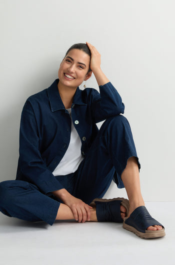 In a navy outfit and Crushed Leather Sliders, this person radiates effortless luxury. Smiling with one hand on their head, the cushioned soles of their sandals provide comfort against the plain backdrop.