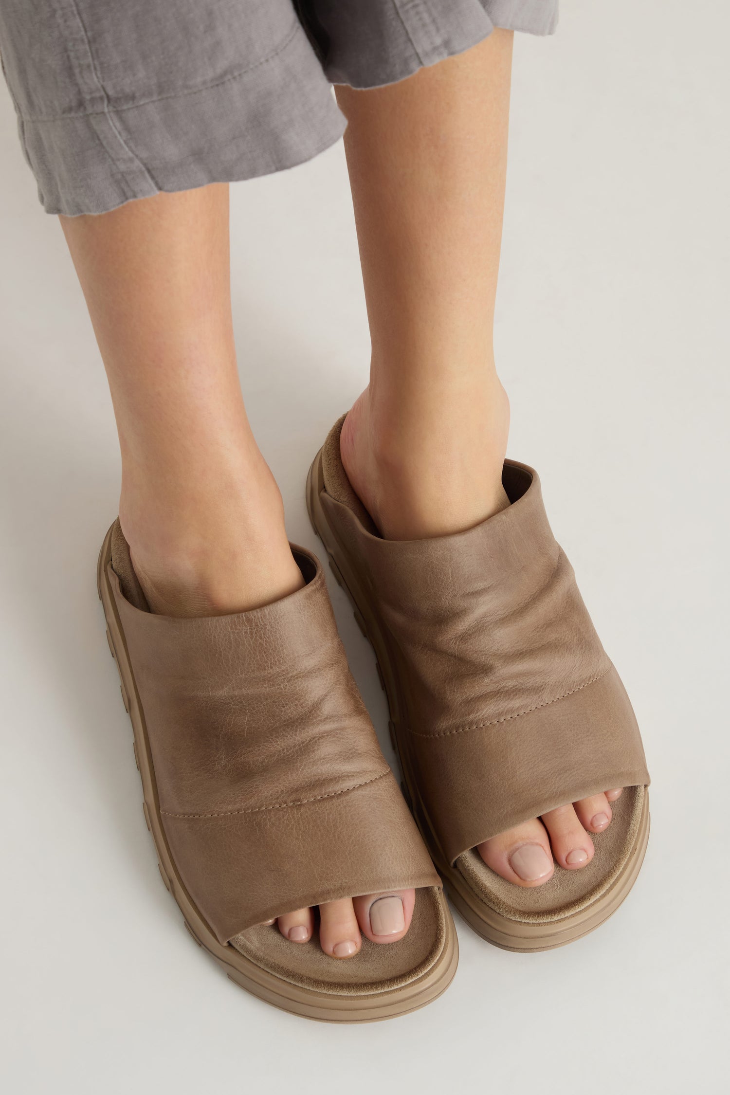 Person wearing Crushed Leather Sliders with thick soles and gray cropped pants, standing on a light surface. Designed for comfort and style.