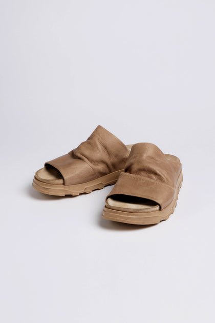 The Crushed Leather Sliders, a pair of tan slip-on sandals with thick soles, are shown on a plain white background. Style LOFI3364 from SS26 offers comfortable versatility for everyday wear.